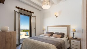 A bedroom with a large bed, a wooden headboard, a nightstand with a lamp, and a view of the outdoors through a sliding door. - El Granadino - Two Bedroom Villa, Sleeps 4 (El Gastor)