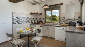 A modern kitchen with a dining area, featuring a glass table, chairs, a built-in oven, and a large window offering a view of greenery. - El Granadino - Two Bedroom Villa, Sleeps 4 (El Gastor)