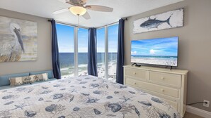 A bedroom with a large bed, a ceiling fan, a TV on a dresser, and a view of the beach through large windows. - The 9th Floor Offers A True Blue View Of The Sparkling Gulf Of Mexico! (pensacola)