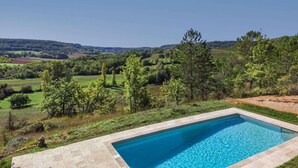 Una piscina rettangolare con superficie d'acqua cristallina azzurra, circondata da un solarium in pietra. Sullo sfondo, un paesaggio verdeggiante con alberi e dolci colline sotto un cielo sereno.