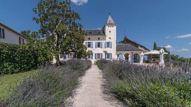 A large, historic building with a tower, surrounded by lavender fields and a paved pathway leading to a covered outdoor seating area.