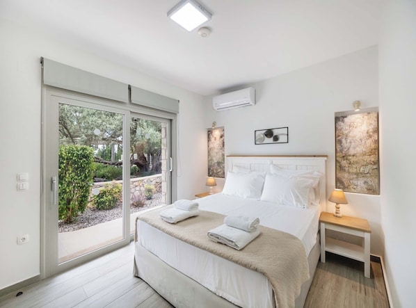 Un dormitorio con cama, dos mesitas de noche, una ventana con vista a la vegetación y un equipo de aire acondicionado.
