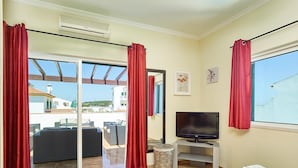 Quarto de hotel com televisão, cama com toalhas, sofá de vime, mesa pequena e janela com cortinas vermelhas.