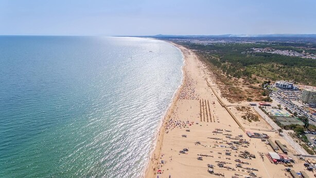 Vista aérea de uma praia com uma extensa faixa de areia, alguns edifícios e céu claro.