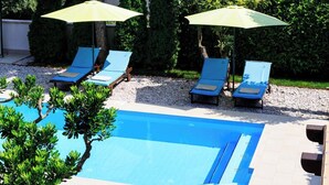 Zona de piscina con sillas de descanso y sombrillas, rodeada de vegetación y montañas al fondo.
