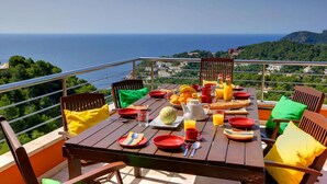 Une table en bois, dressée pour le petit-déjeuner, avec vue sur la mer et la verdure.