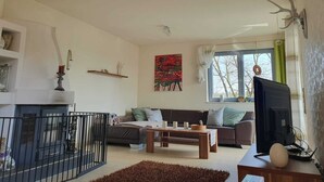 A living room with a sofa, coffee table, and TV. There is a fireplace, a painting on the wall, and a window with curtains. - Relaxation oasis in the heart of Rheinhessen, Haus Weinbergsblick (Armsheim)