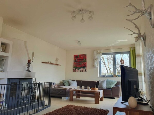 A living room with a sofa, coffee table, and TV. There is a fireplace, a painting on the wall, and a window with curtains.