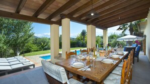 An outdoor dining area with a wooden table set for six, surrounded by cushioned chairs. A pool is visible in the background, and the area is covered by a wooden pergola. - Ca'n Jaume Barretes - Four Bedroom Villa, Sleeps 8 (Mallorca)