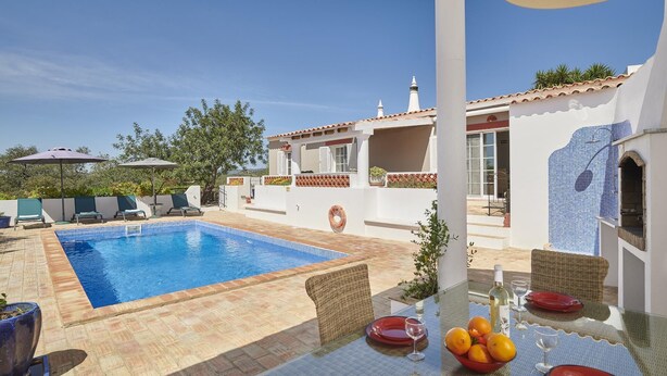 Un espace aménagé au bord de la piscine, avec une piscine à l’eau d’un bleu limpide, des chaises longues et une table à manger dressée avec des assiettes, des verres et une bouteille.