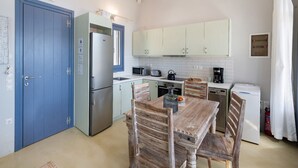 A compact kitchen with a refrigerator, microwave, kettle, and coffee maker. There is a wooden dining table with chairs.