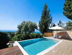 A swimming pool with a clear view of the sea, surrounded by trees and a stone wall.