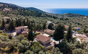 A residential area with houses surrounded by dense trees, a clear view of the sea, and a hilly landscape in the background.