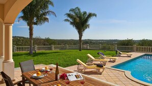 Un espace repas aménagé au bord de la piscine, avec une table pour deux, entouré de chaises longues, d’une piscine et de palmiers.