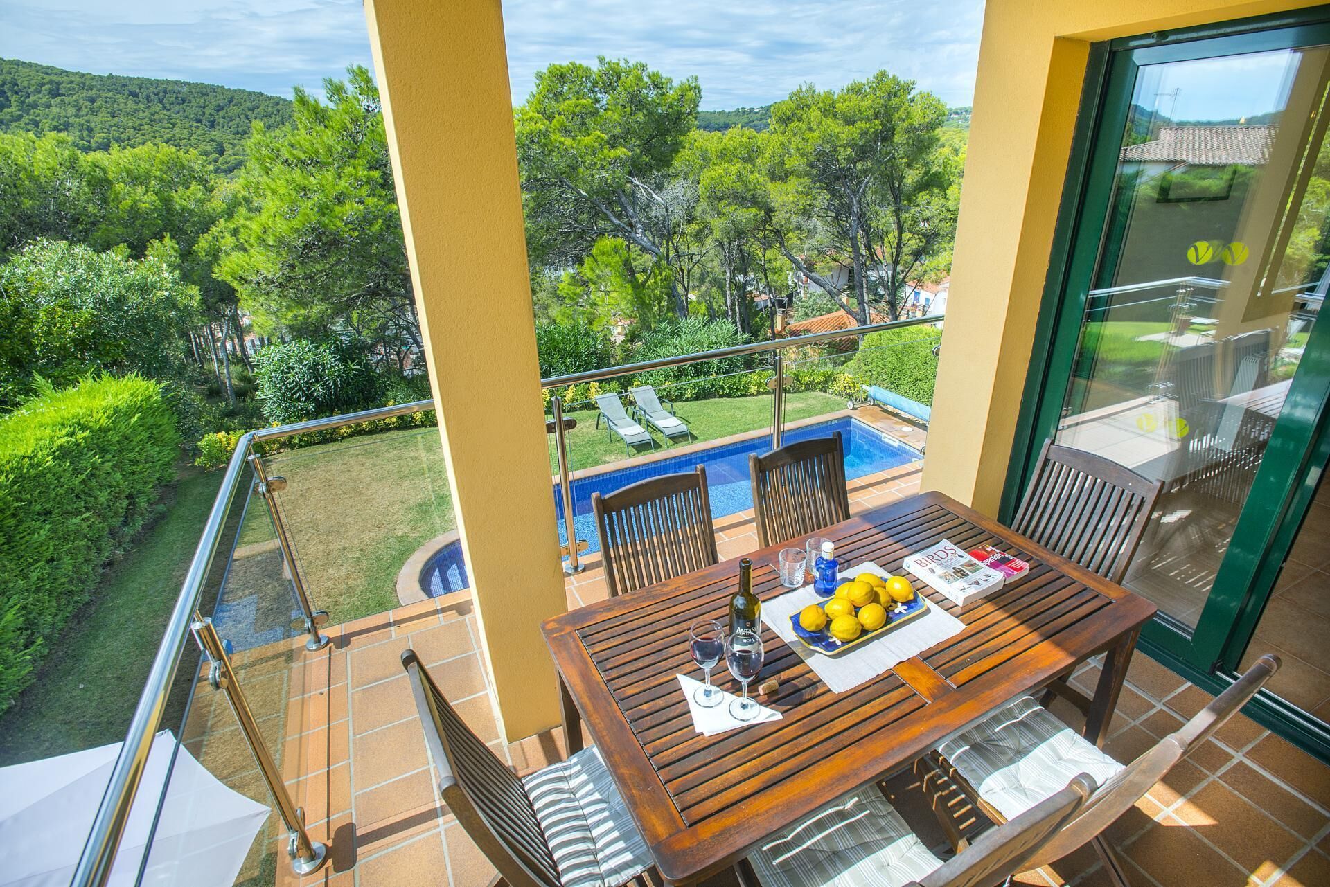 A balcony with a wooden table, chairs, and a view of a swimming pool and greenery.