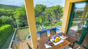 En balkon med et træbord, stole og udsigt til en swimmingpool og grønne områder.