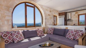 A cozy living area with a stone wall, a large arched window, and a modern kitchen in the background. - Villa Kourouni - Three Bedroom Villa, Sleeps 6 (Crete)