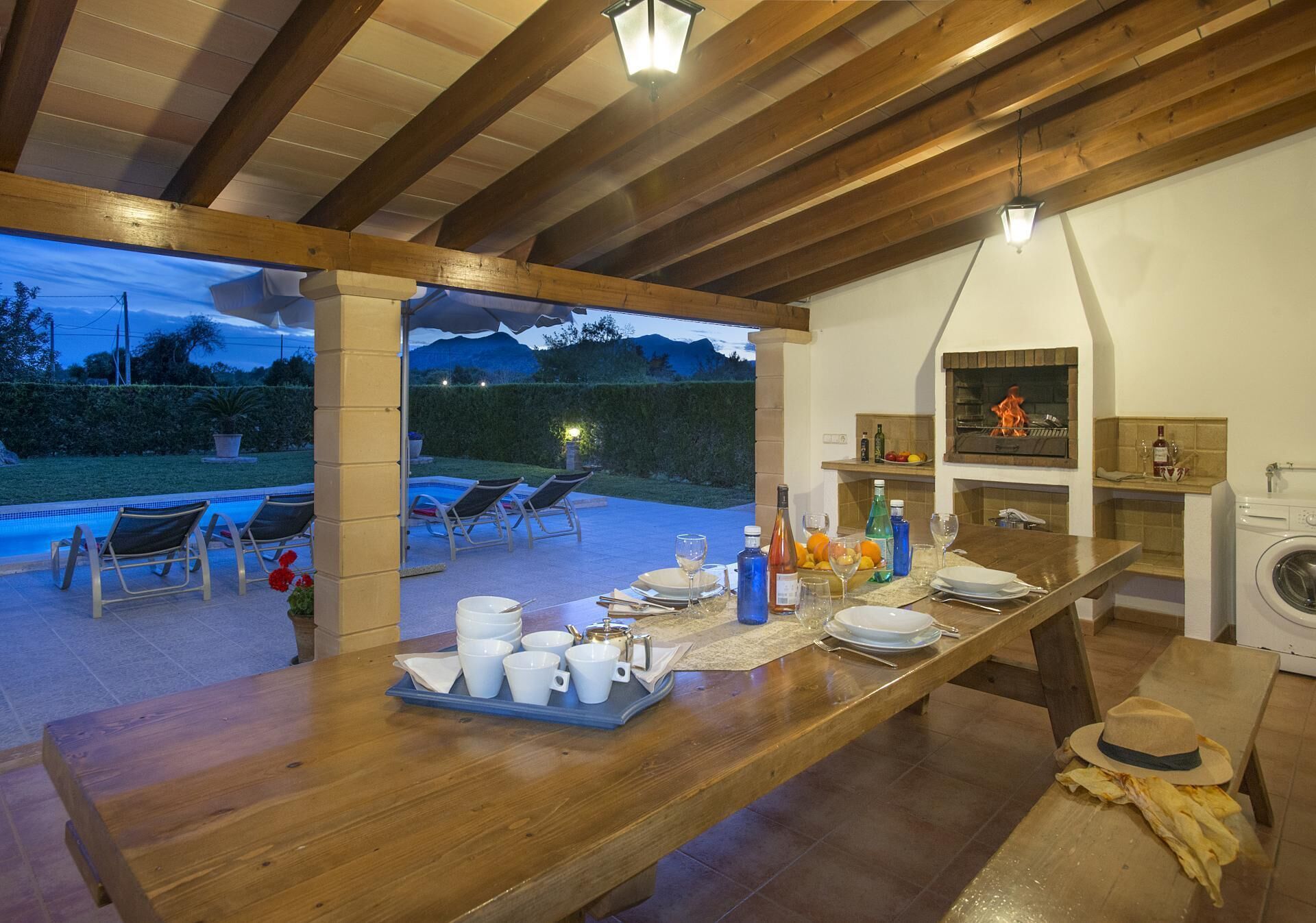 Un coin repas avec une table en bois dressée pour le repas, une cuisine équipée d’une cuisinière et un espace détente au bord de la piscine avec des chaises.