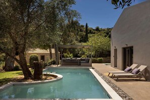 Une piscine dotée d’une pergola et d’un espace détente, entourée d’arbres et d’un bâtiment.
