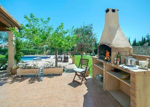 Een terras buiten met een stenen barbecue, een zwembad en een pergola.