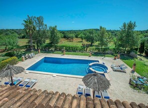 Une piscine rectangulaire avec un jacuzzi, entourée de chaises longues et de parasols, le tout se fondant dans un décor de verdure et sous un ciel dégagé.