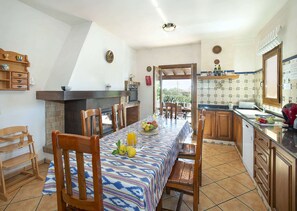 Un espace repas comprenant une longue table, des chaises, une cheminée et un coin cuisine avec des placards et un dosseret carrelé.