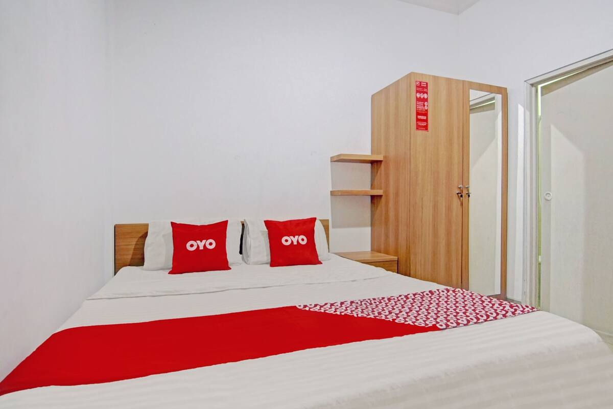 Una habitación de hotel con una cama, dos almohadas rojas con la marca "OYO", un armario de madera y un plaid rojo y blanco.