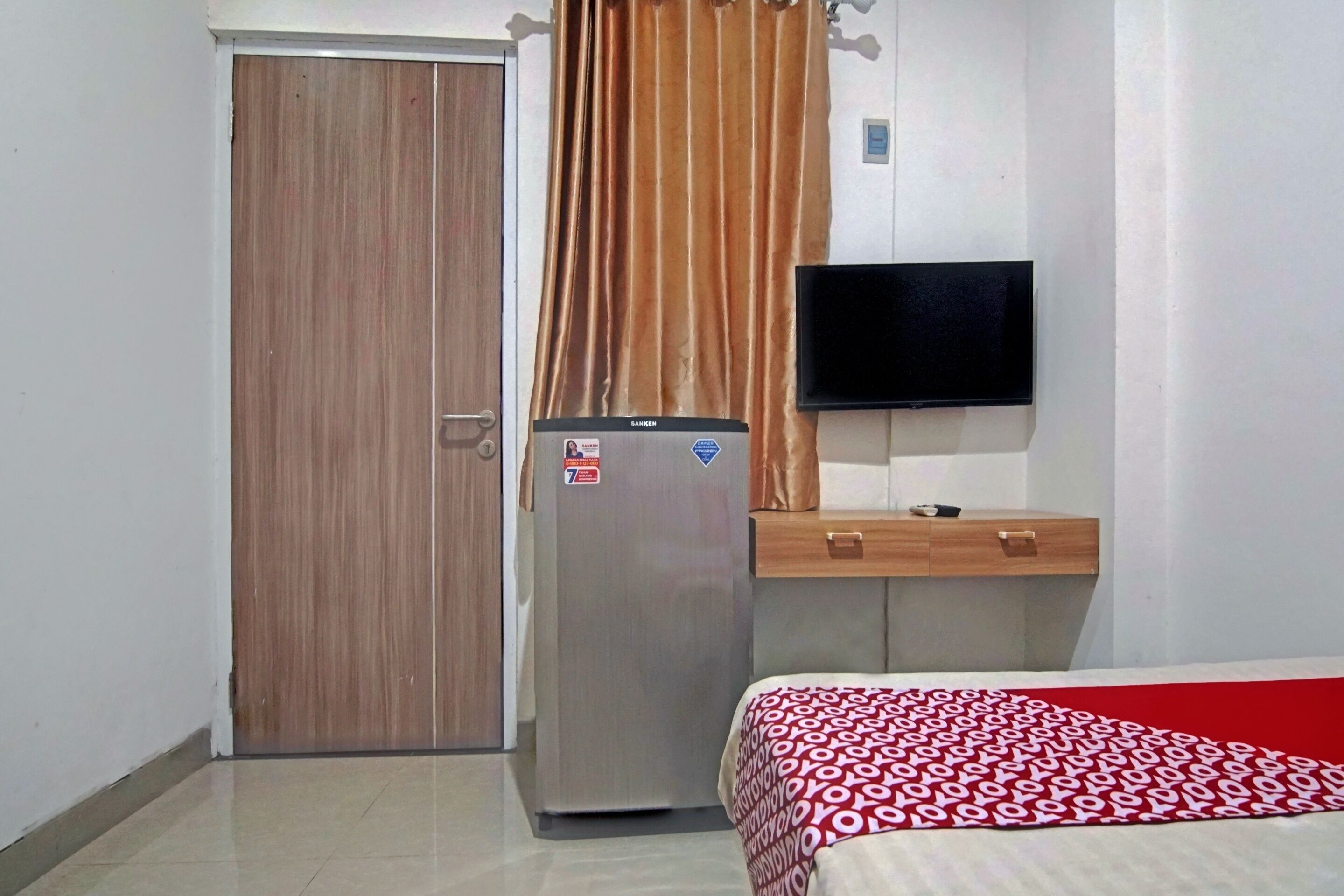A hotel room with a wooden wardrobe, a refrigerator, a television mounted on the wall, and a bed with a patterned bedspread.