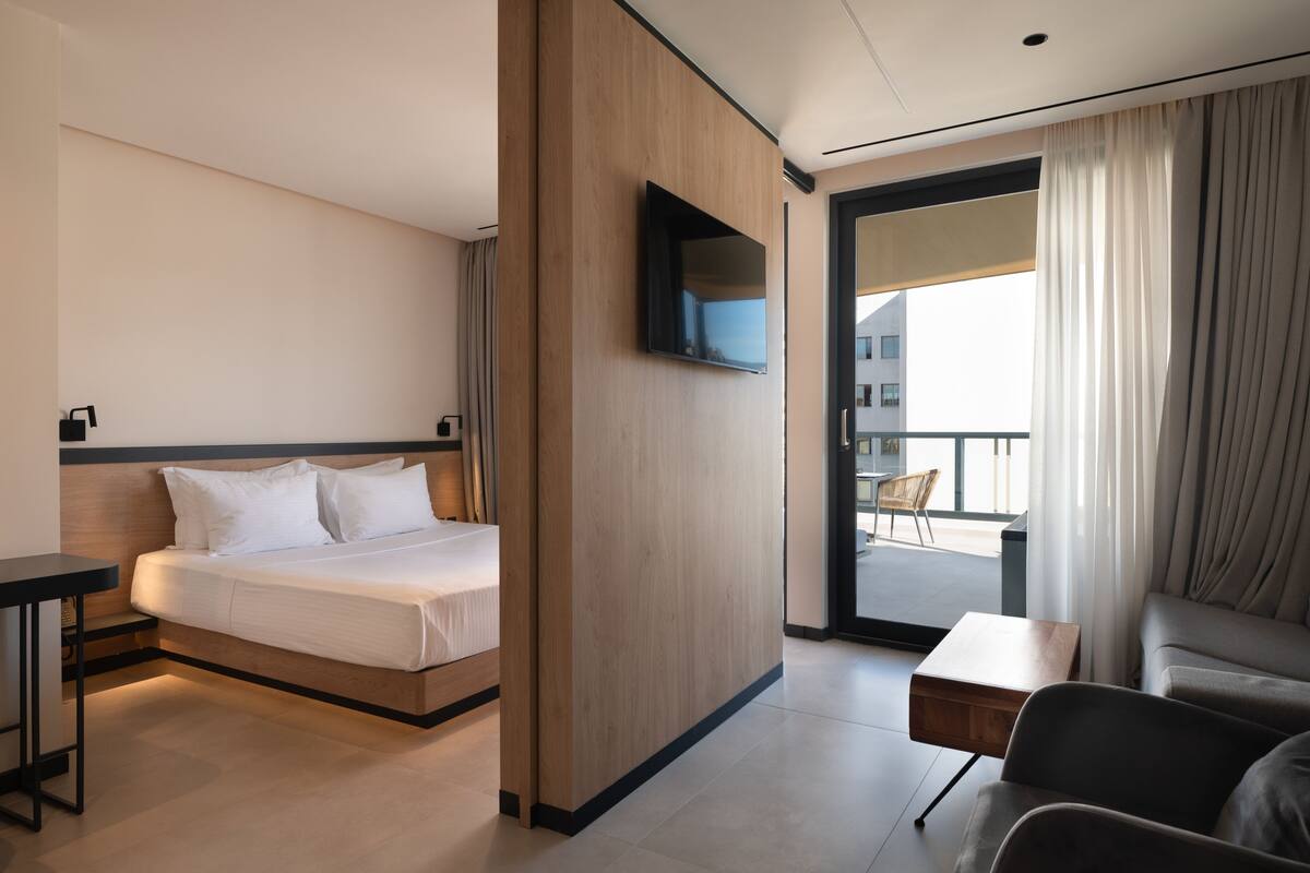 Una habitación de hotel moderna con una cama, un televisor en la pared, un balcón con una silla y un sofá.