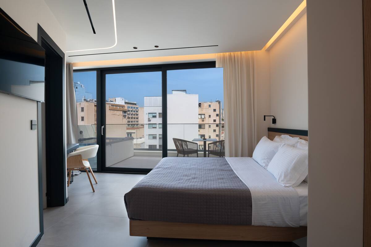 Habitación de hotel moderna con una cama grande, un escritorio y una silla, un balcón con vistas a la ciudad y un televisor montado en la pared.