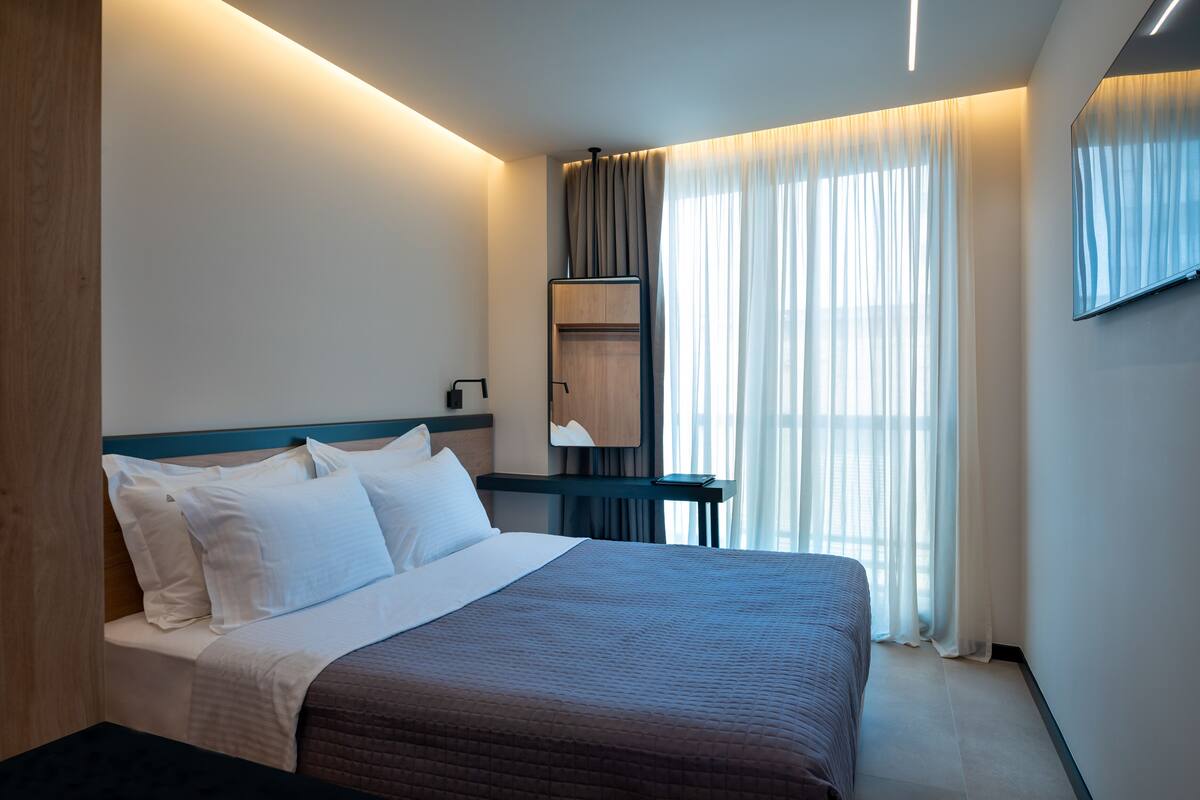 Una habitación de hotel moderna con una cama grande, una mesita de noche, un espejo y un ventanal con cortinas.