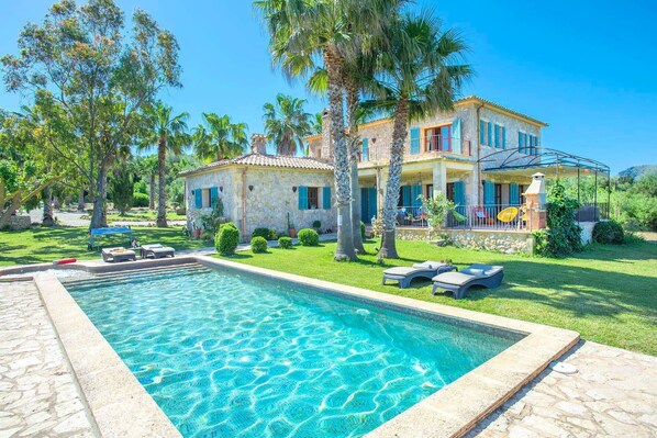 A two-story stone house with a swimming pool, surrounded by lush greenery and palm trees.