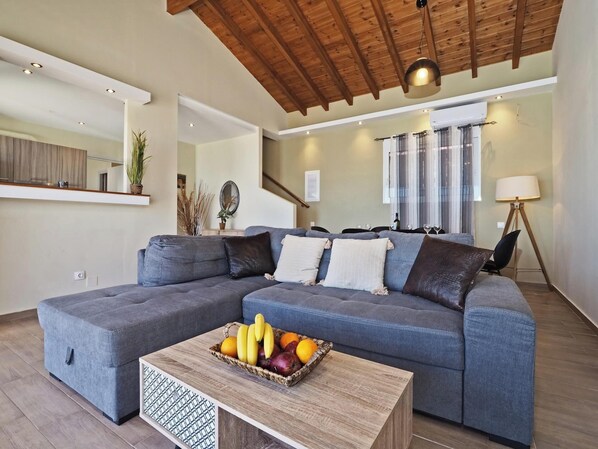 Ein modernes Wohnzimmer mit einem grauen Ecksofa, einem Couchtisch mit Obst und einer Holzbalkendecke.