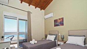 A hotel room with two beds, a desk, and a chair. There is a view of the sea through a large window. - Sun Mare - Three Bedroom Villa, Sleeps 6 (Corfu)