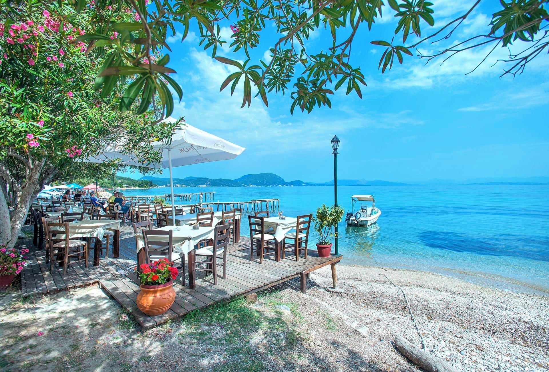 An outdoor dining area with tables and chairs, overlooking a calm sea with boats.