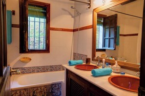 Une salle de bain avec une baignoire, deux lavabos, un grand miroir et une fenêtre donnant sur un paysage verdoyant.