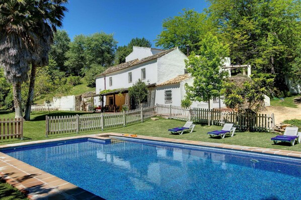 Une piscine entourée d’une clôture blanche, une maison blanche avec un toit en tuiles et une végétation luxuriante.