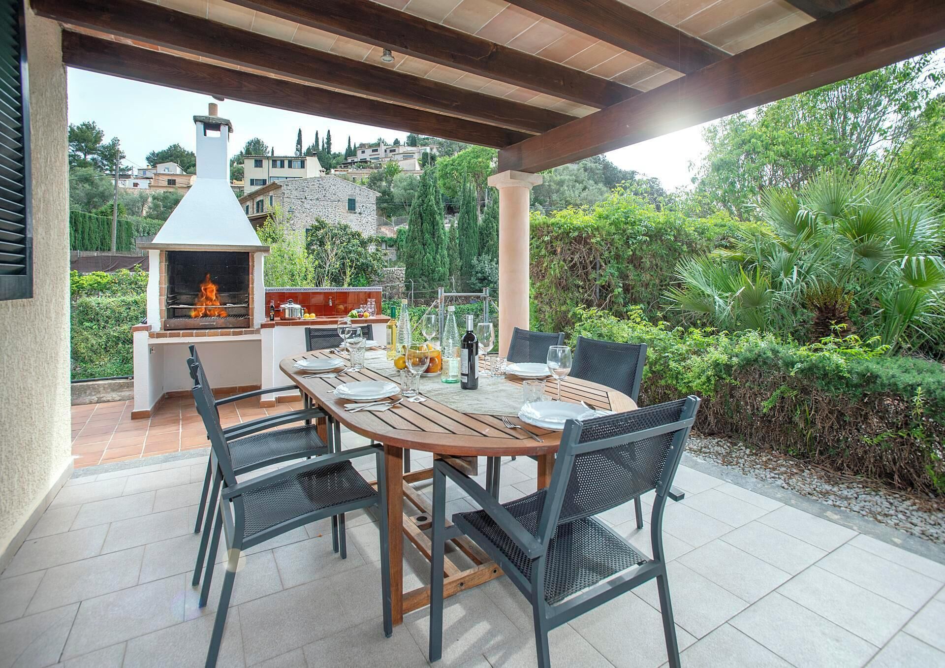 An outdoor patio with a wooden table set for dining, surrounded by chairs, and a barbecue grill in the background.