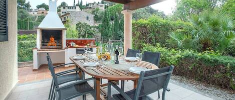 An outdoor patio with a wooden table set for dining, surrounded by chairs, and a barbecue grill in the background.