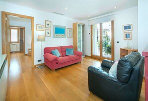 A spacious living room with a red sofa, a black leather chair, and a wooden floor. There are framed pictures on the walls and a window with a view of greenery outside.