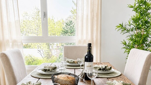 A dining table set with plates, glasses, and a bottle, surrounded by chairs and a view of greenery.