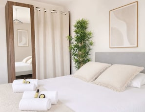 A hotel room with a bed, white towels, a mirror, a plant, and a framed abstract artwork.