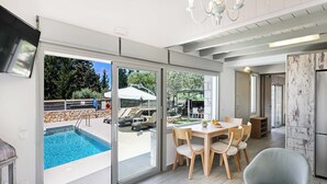Une cuisine moderne dotée d’un four encastré, un coin repas avec une table et des chaises, et une grande porte-fenêtre coulissante donnant sur une piscine extérieure.