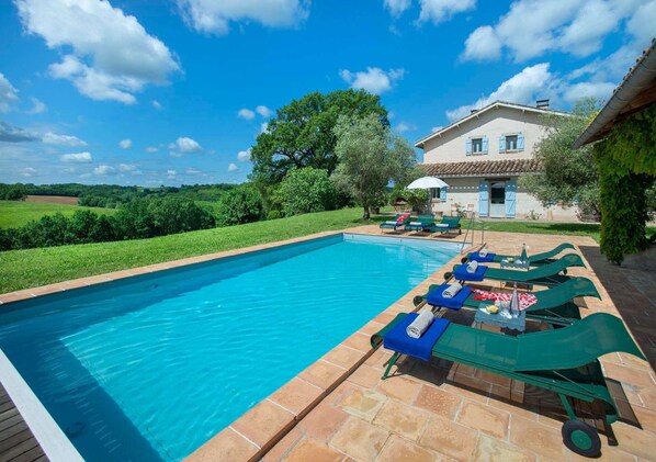 A swimming pool with lounge chairs and a table, surrounded by a garden and a house.