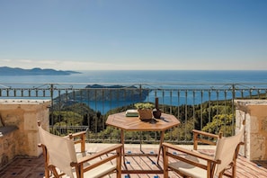 A balcony with a table and chairs, overlooking a scenic view of the sea and hills.
