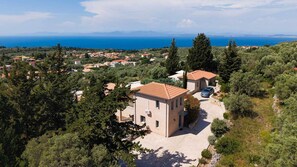 A Mediterranean-style villa with a paved driveway, surrounded by dense greenery and overlooking a coastal landscape.