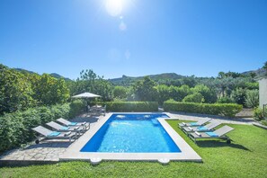 Una piscina rectangular con sillones de descanso y una mesa, rodeada de césped y setos cuidados, con vistas despejadas a colinas al fondo.