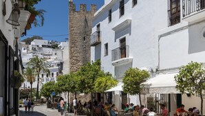 A narrow street lined with white buildings, some with balconies and others with large windows. There are outdoor seating areas with tables and chairs, and a few people are visible. A tall stone tower is visible in the background. - Casa Bellota - One Bedroom Villa, Sleeps 2 (Vejer de la Frontera)