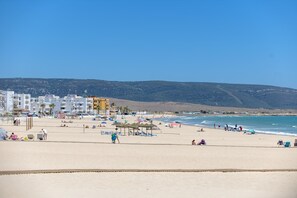 Una playa con edificios al fondo, cielos despejados y gente disfrutando.