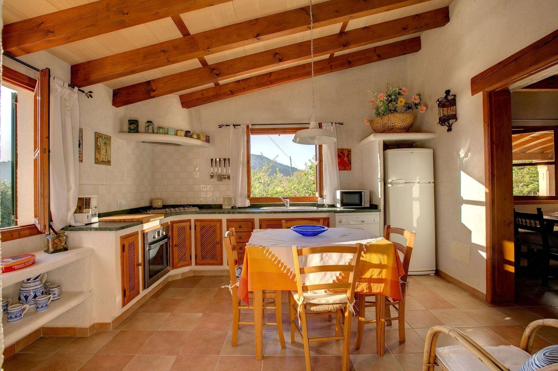 A cozy kitchen with wooden beams, a dining table with chairs, a refrigerator, and a window with a view of greenery.